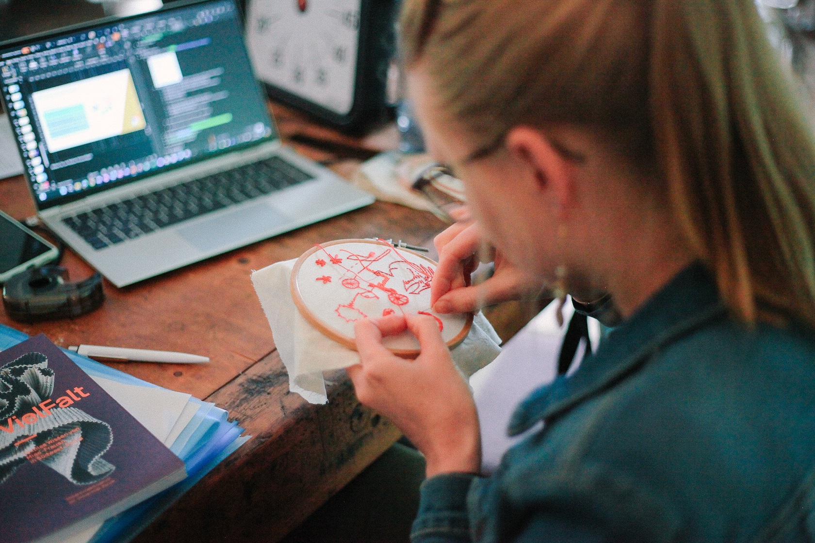 Eine Frau stickt an einem Handstickrahmen; im Hintergrund ist ein Laptop zu sehen.